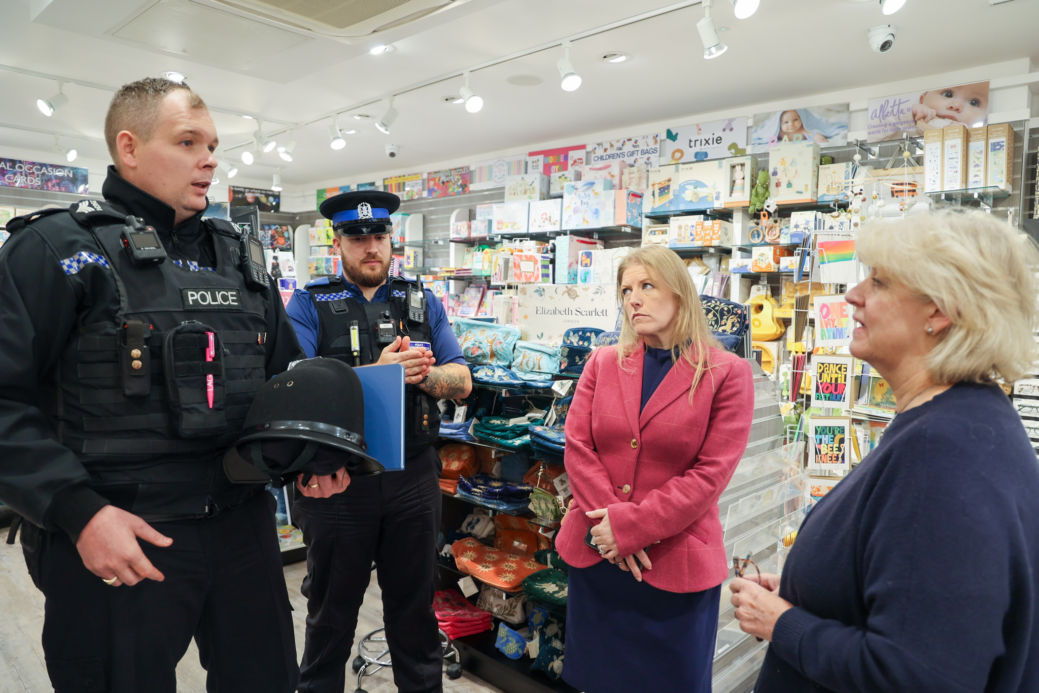 Two police officers speak with the Police and Crime Commissioner and a retailer inside a gift and stationery shop. The group stands among brightly lit shelves filled with cards, toys and accessories, discussing retail crime and local issues.